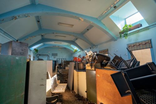 A cluttered room filled with discarded furniture and appliances, under a blue ceiling with sunlight filtering through windows.