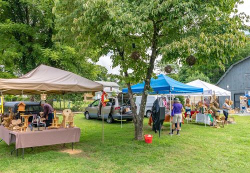 A vibrant outdoor market with tents, vendors, and shoppers among trees and grassy areas.