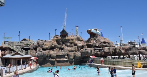 A water park scene with a rocky structure, a boat, and people swimming in a pool under a clear blue sky.