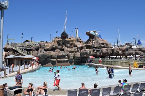 A busy water park scene with a wave pool, rocky structures, and people enjoying the water on a sunny day.