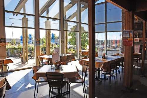 Bright restaurant interior with wooden tables, large windows, and outdoor seating visible through the glass.