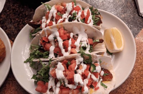 Three tacos filled with lettuce, tomatoes, and drizzled with sauce, served with a lemon wedge on a plate.