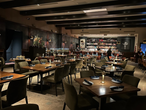 A modern restaurant interior with wooden tables, plush chairs, and a well-stocked bar in the background.