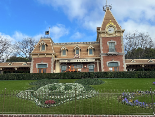 Disneyland entrance with a clock tower, surrounded by flower beds shaped like Mickey Mouse. Clear blue sky above.