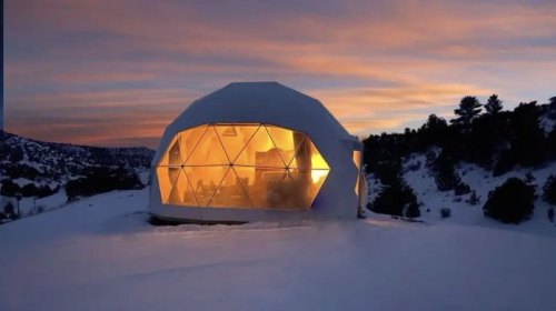 A geodesic dome home illuminated at sunset, surrounded by snow-covered landscape and mountains.