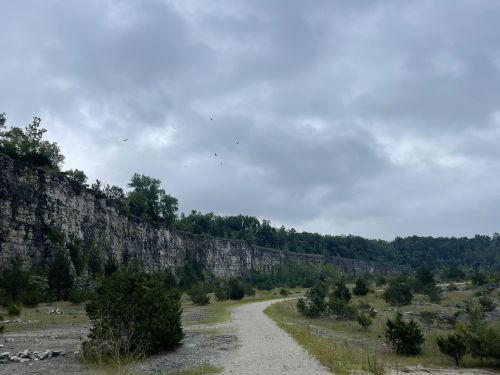 A gravel path leads through a rocky landscape under a cloudy sky, with trees and a cliff in the background.