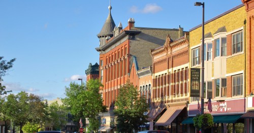 Historic buildings line a street under a clear blue sky, with trees and shops adding to the charming atmosphere.