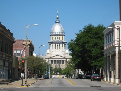 View of a wide street leading to a large domed state capitol building, flanked by trees and historic buildings.