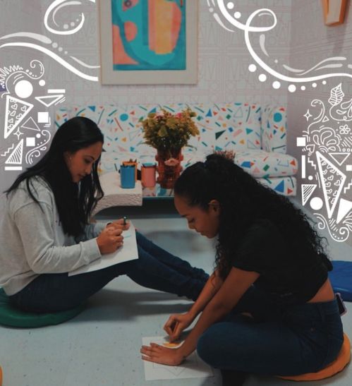 Two women sit on the floor, drawing and writing, surrounded by colorful decor and a floral arrangement.