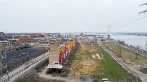 Aerial view of an industrial area with colorful murals on a building, power lines, and a river in the background.