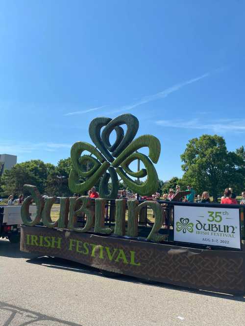 A large green shamrock sign for the Dublin Irish Festival, with a crowd in the background on a sunny day.