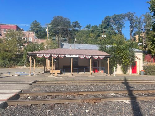 A small train station with a covered platform, decorated with bunting, surrounded by trees and buildings under a clear blue sky.