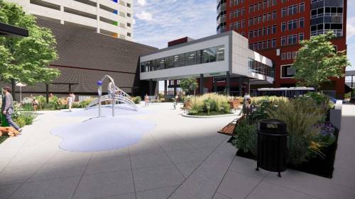 A modern urban plaza with greenery, a splash pad, and people enjoying the space near contemporary buildings.