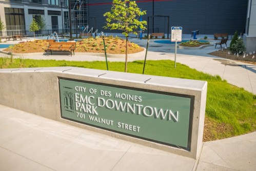 Sign for EMC Downtown Park in Des Moines, featuring green grass, benches, and a modern urban landscape.