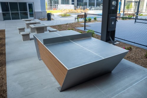 A modern outdoor table with a sleek design, surrounded by stone benches and greenery in a landscaped area.