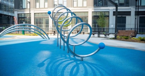 A modern playground with blue rubber flooring and metal climbing structures in a sunny urban setting.