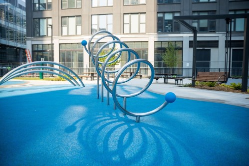 Playground with blue rubber surface and metal climbing structure, surrounded by modern buildings and greenery.