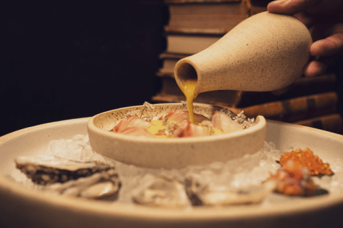 A hand pours sauce over a dish of seafood on ice, surrounded by oysters and garnishes, with books in the background.