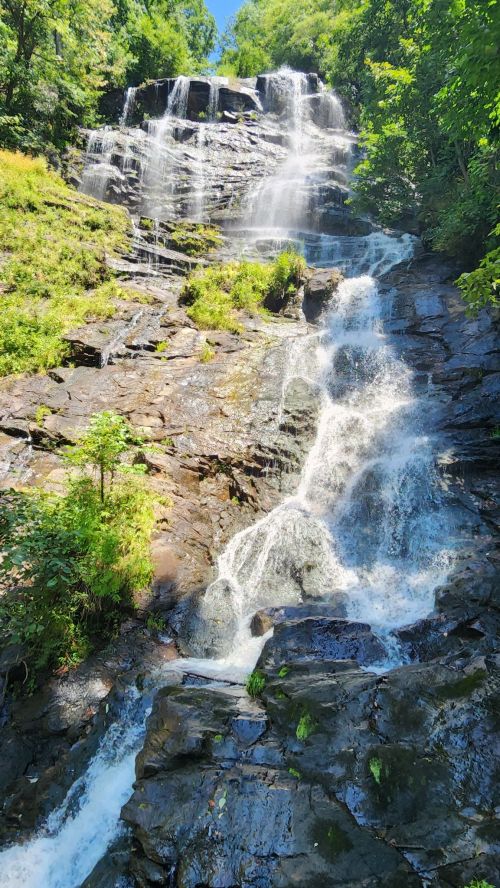 A cascading waterfall flows down rocky terrain, surrounded by lush greenery and bright blue skies.