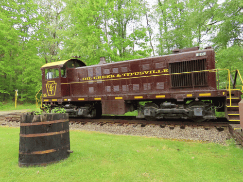 A vintage train from Oil Creek & Titusville Railroad parked on tracks surrounded by lush green trees.