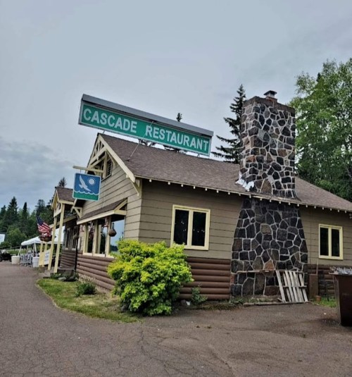 A rustic building with a stone chimney and a sign reading "Cascade Restaurant," surrounded by trees and greenery.