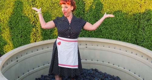 A woman in a polka dot dress and apron stands playfully in a grape-filled vat, surrounded by greenery.