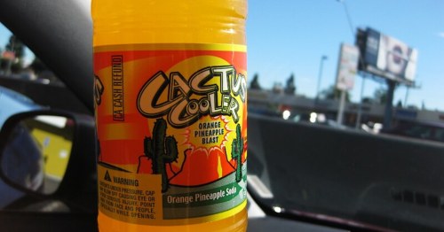 A close-up of a Cactus Cooler orange pineapple soda bottle on a car dashboard, with a billboard in the background.