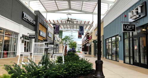 A view of a shopping mall corridor with stores on either side and greenery in the center under a glass roof.