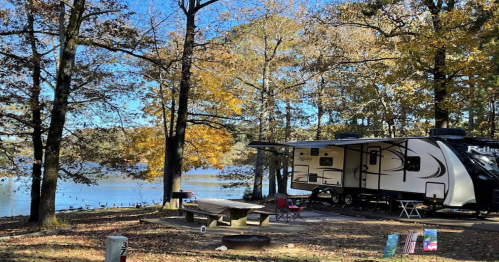 A camper parked by a serene lake, surrounded by autumn trees with colorful leaves and a picnic area nearby.