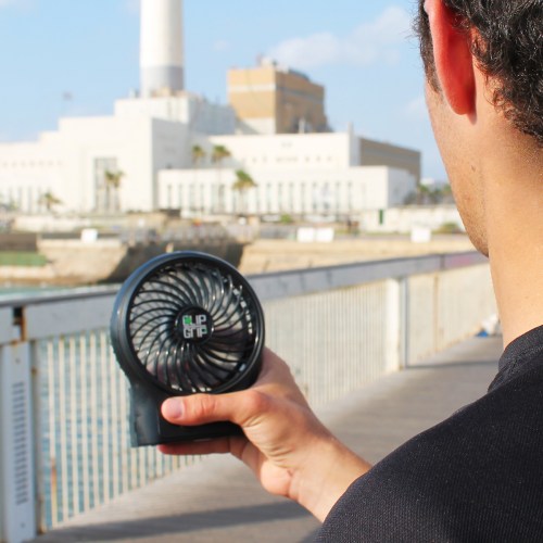 A person holds a portable fan while standing on a boardwalk near a waterfront and a large building in the background.