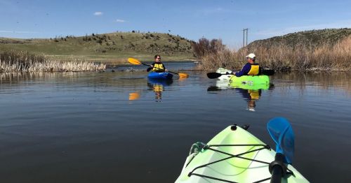 Two kayakers paddle on a calm lake surrounded by reeds and hills under a clear blue sky.