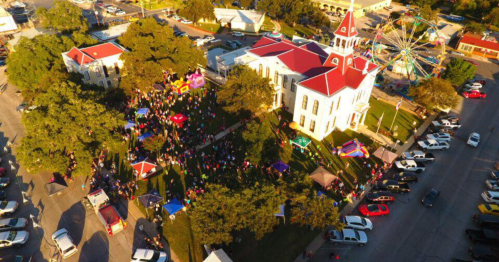 Aerial view of a festive gathering with a carnival, colorful tents, and a historic building surrounded by people and cars.