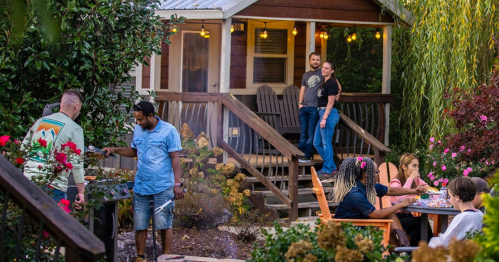 A group of people enjoying a backyard barbecue near a cozy cabin, surrounded by greenery and flowers.