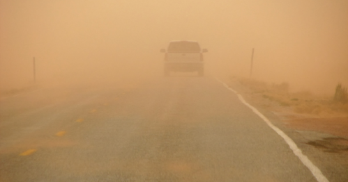 A white vehicle drives down a deserted road shrouded in dust and fog, creating a hazy, atmospheric scene.