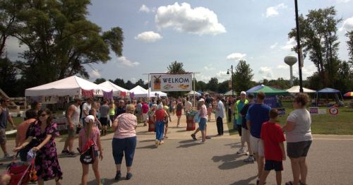 A lively outdoor festival scene with people walking among vendor tents under a sunny sky. A "WELKOM" sign is visible.