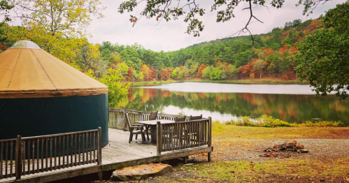 A cozy yurt by a serene lake, surrounded by colorful autumn trees and a wooden deck with a table and chairs.