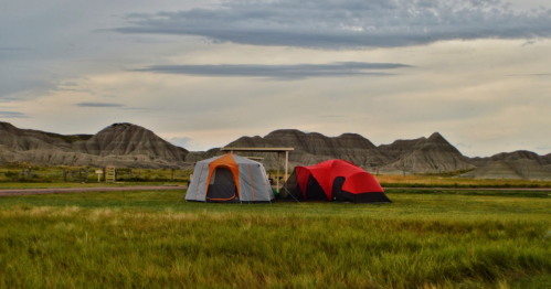 Two tents, one orange and one red, set up on grassy land with rugged hills in the background under a cloudy sky.