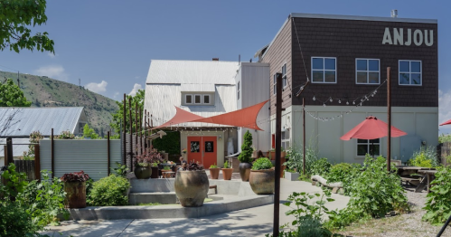 A modern building with a sign reading "ANJOU," surrounded by greenery and outdoor seating under red umbrellas.