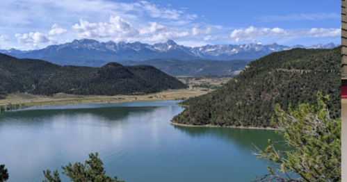 A serene lake surrounded by mountains under a blue sky with scattered clouds. Lush greenery frames the scene.