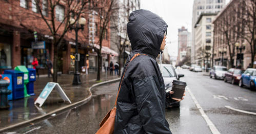 A person in a raincoat holds a coffee cup while walking down a wet city street lined with buildings and parked cars.