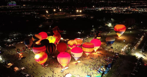 Aerial view of a vibrant hot air balloon festival at night, with colorful balloons illuminated and crowds below.
