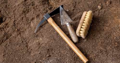 Three gardening tools—a pick, a small hoe, and a brush—resting on brown soil.