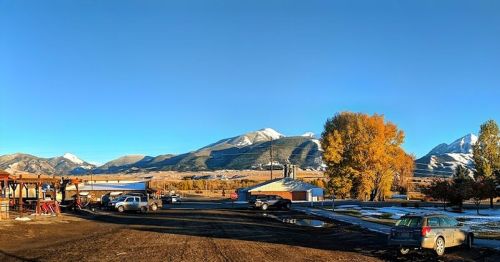 A scenic view of mountains and autumn trees, with cars parked in a rural area under a clear blue sky.