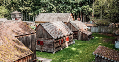 Aerial view of rustic wooden cabins surrounded by trees and green grass in a historical village setting.