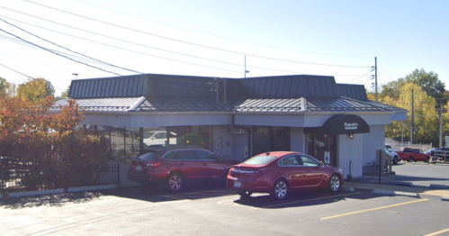 A small restaurant building with a black roof, surrounded by parked cars and trees in the background.