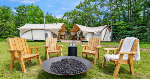A cozy outdoor campsite with wooden chairs around a fire pit, featuring glamping tents in a lush green setting.