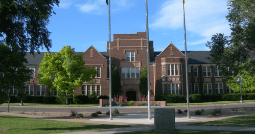Historic brick school building with green trees and flags in front, under a clear blue sky.