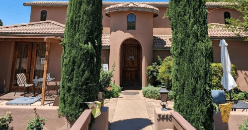 A charming adobe-style house with tall cypress trees, a wooden door, and a welcoming pathway.