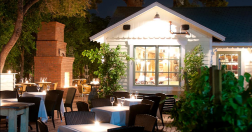 A cozy outdoor restaurant at night, featuring tables with white tablecloths and a charming white building in the background.