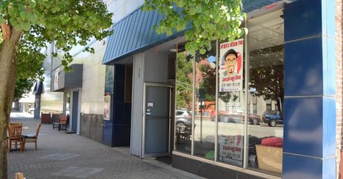 A storefront with large windows, featuring a sign and trees lining the sidewalk on a sunny day.
